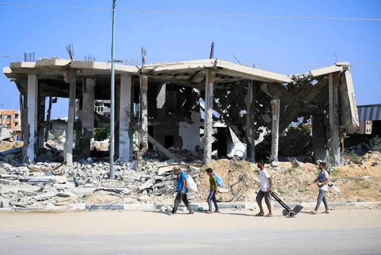 Palestinos deslocados carregam pertences enquanto caminham em frente a um prédio destruído em Deir el-Balah, na região central da Faixa de Gaza (Eyad Baba/AFP)